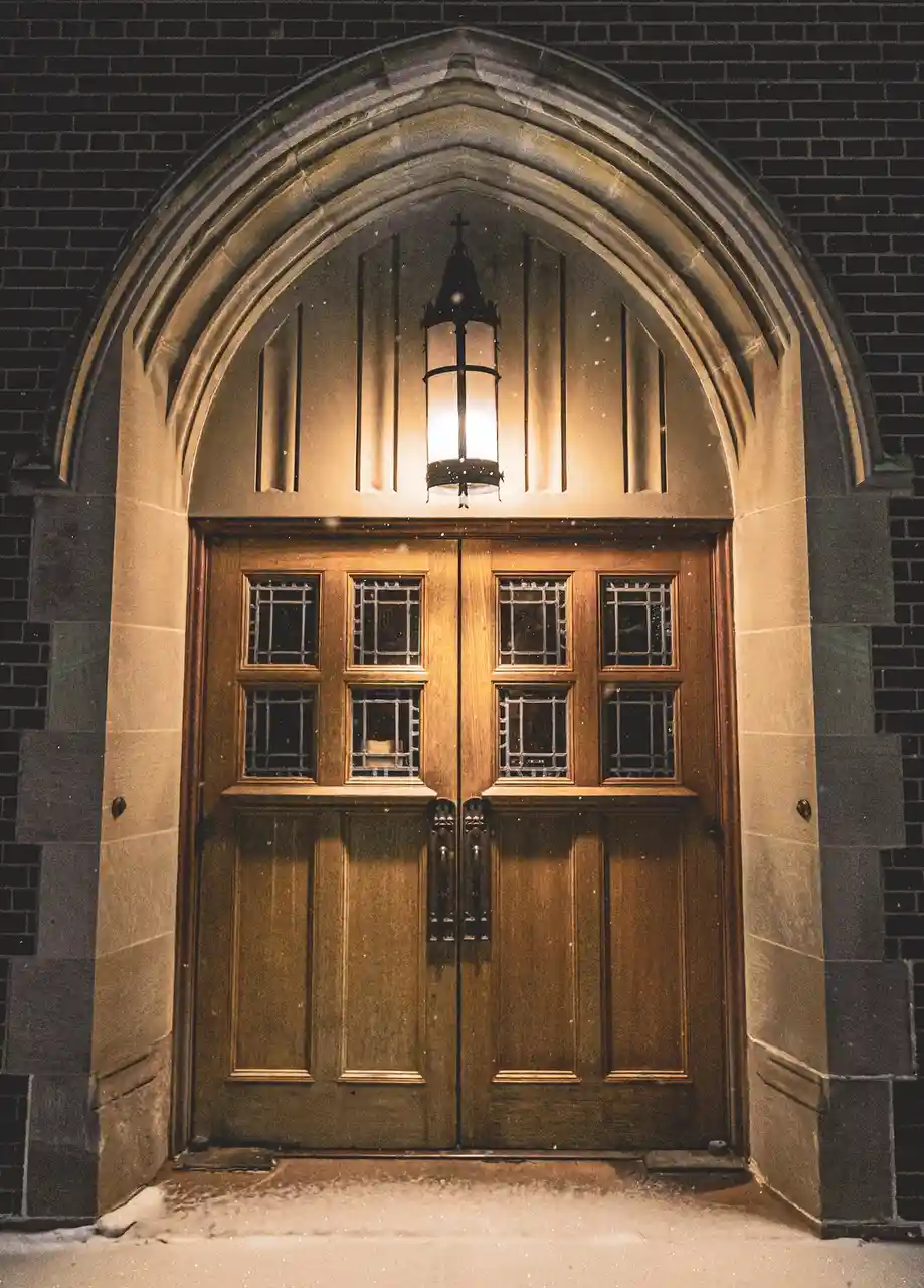 A warmly lit wooden church door with stained glass windows is framed by a stone archway under falling snow. A lantern hangs…