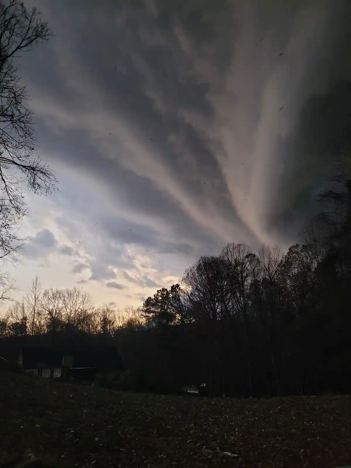 A dark, ominous sky with dramatic cloud formations looms over a silhouette of bare trees and a house at dusk. Numerous small…