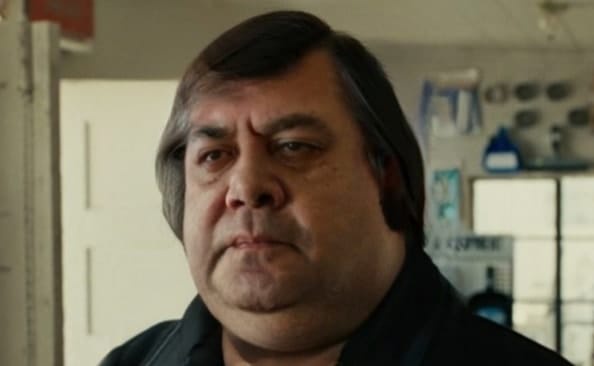 A man with a stern, unimpressed facial expression stares directly at the camera. He has dark hair, a heavy brow, and a…