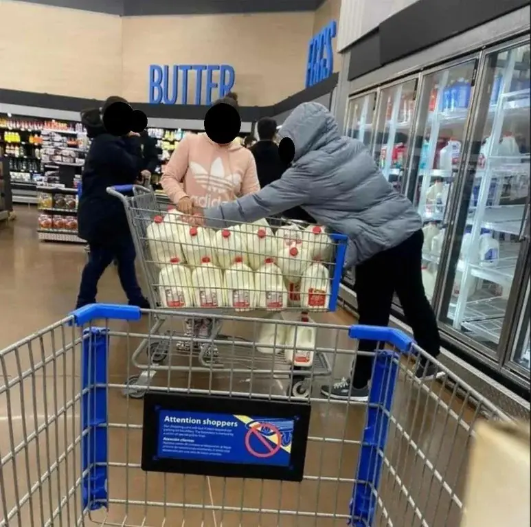 A person in a grey hoodie is pushing a shopping cart overflowing with milk jugs in a grocery store aisle labeled 'BUTTER'…