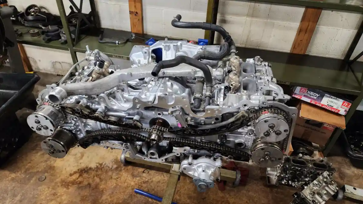 A disassembled car engine block sits on a workbench in a garage, surrounded by tools and parts. The engine is partially…