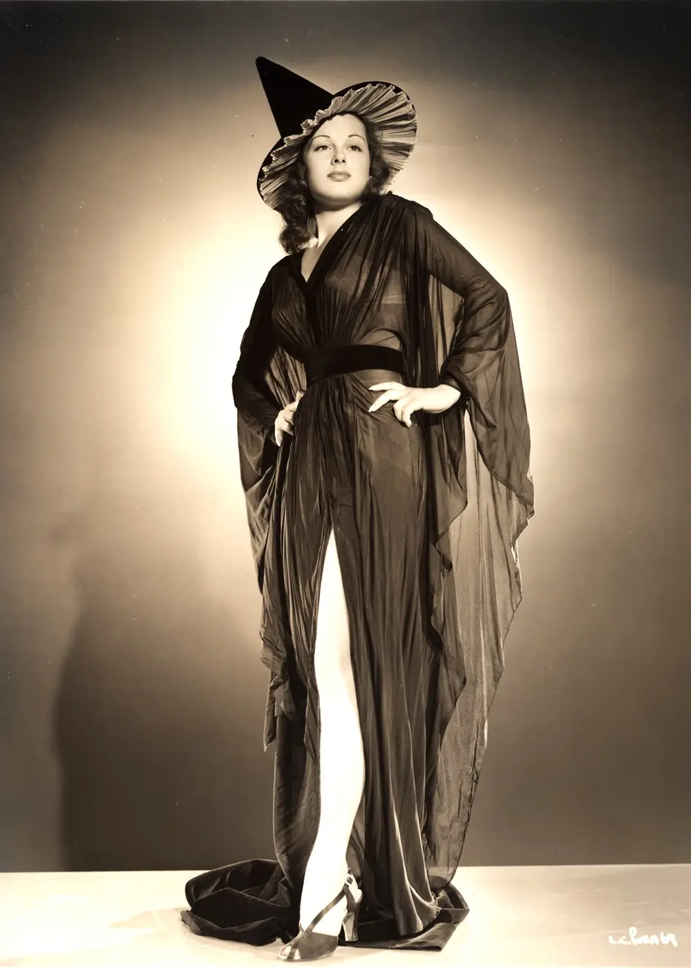 A vintage black-and-white studio portrait shows a woman posing confidently in a dramatic, sheer black gown with a high slit…