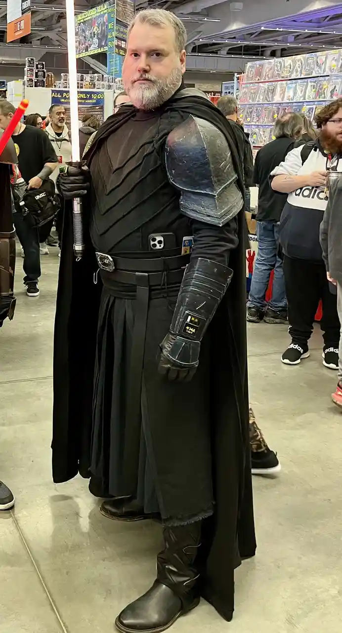 A man with a gray beard and mustache dressed as a Star Wars character in a black robe, armor, and boots, holding a…