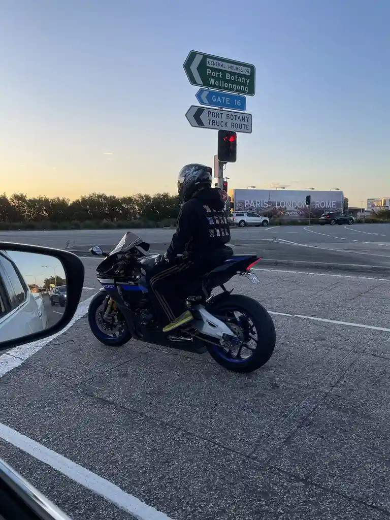 A motorcyclist in full gear waits at a red light at an intersection with road signs pointing to Port Botany and Wollongong…