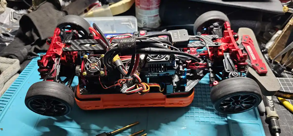 A close-up photo of a highly modified red and black radio-controlled car with exposed wiring, a visible motor labeled…