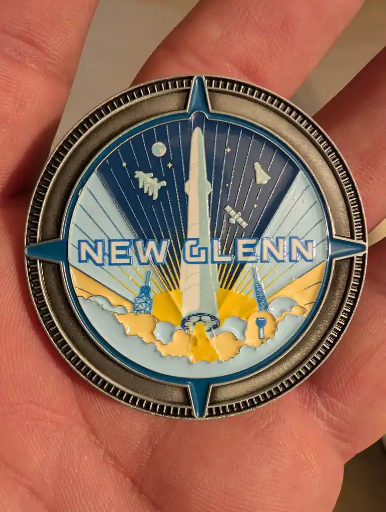 A hand holds a circular, silver-colored enamel pin featuring a stylized rocket launch scene with the text 'NEW GLENN' across…