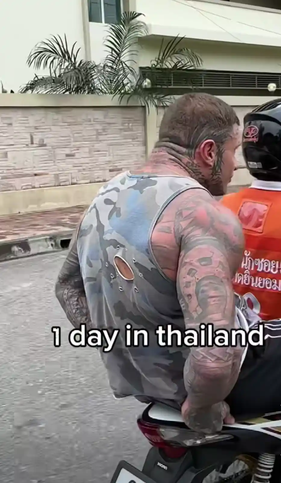 A heavily tattooed man with a camouflage-patterned tank top, featuring a large circular cutout on the back, is riding a…