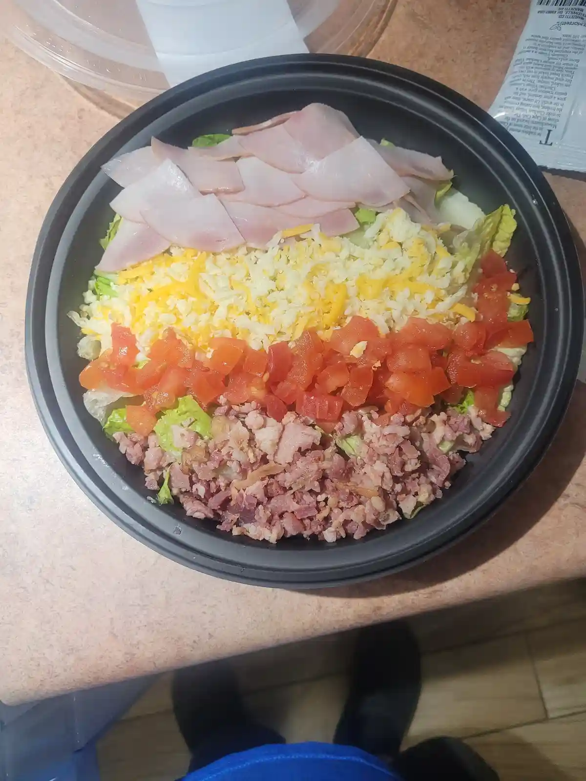 A top-down view of a black plastic bowl filled with a colorful, layered salad featuring bacon, diced tomatoes, shredded…