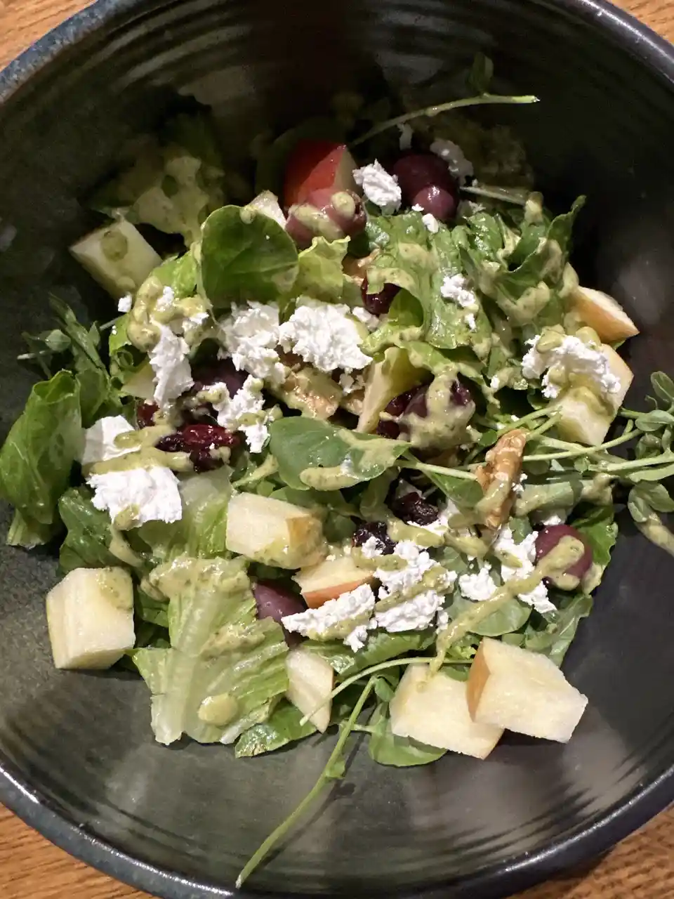 A close-up photo of a dark bowl filled with a colorful salad featuring mixed greens, diced apples, red grapes, crumbled…