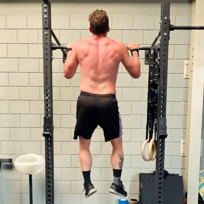 A shirtless man with a muscular back is hanging from a pull-up bar in a gym, viewed from behind. He wears black shorts…
