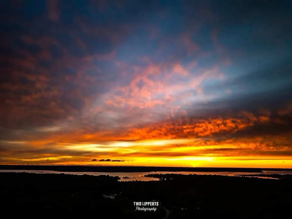 A dramatic sunset photograph showing fiery orange and red clouds against a dark blue sky over a silhouetted landscape with…