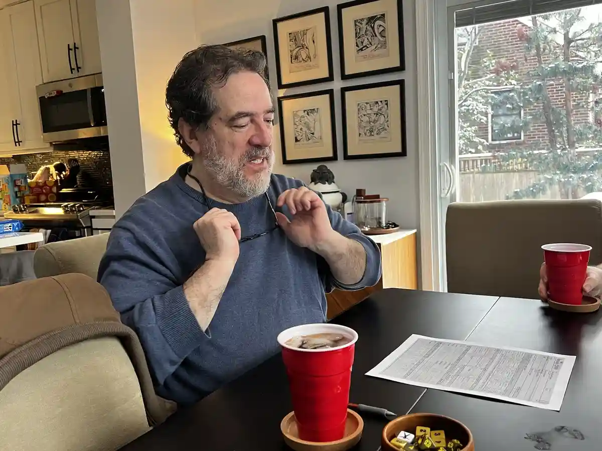 A middle-aged man with a beard and glasses, wearing a blue sweater, sits at a dining table in a home, holding his glasses as…