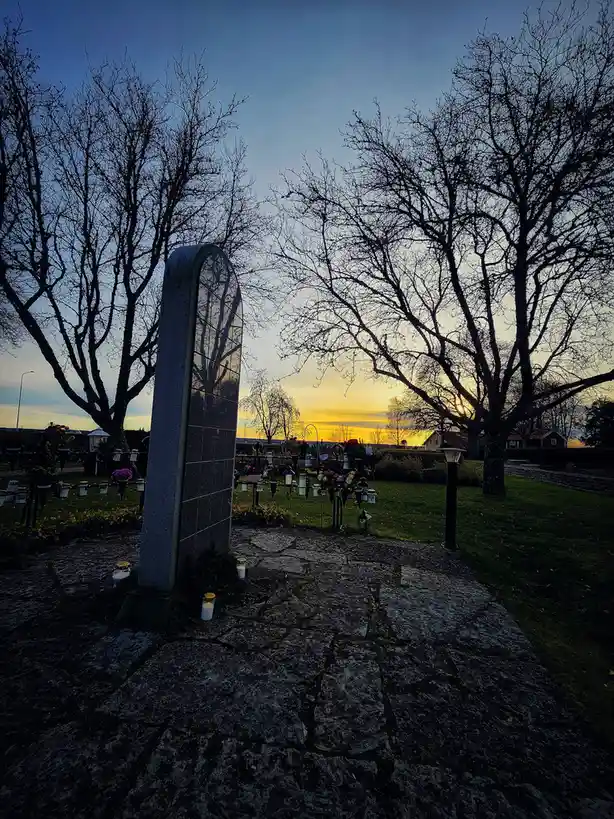 A serene cemetery scene at dusk, featuring a tall, arched gravestone with candles placed at its base, surrounded by bare…
