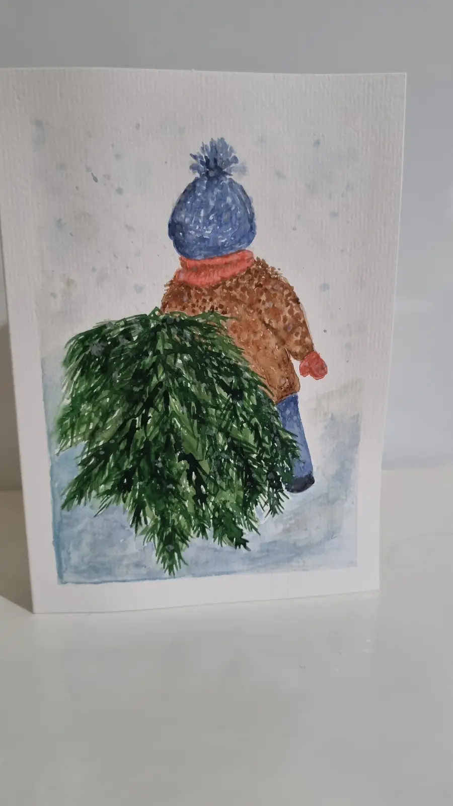 A watercolor painting on a card shows a child from behind, wearing a blue pom-pom hat, orange scarf, brown coat, and blue…