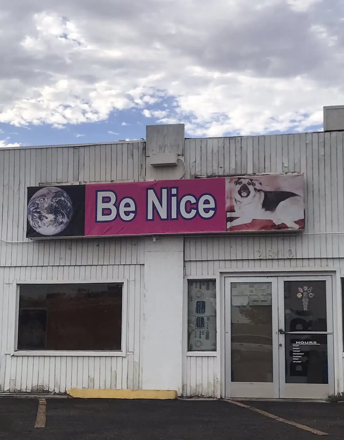 A weathered white building with a large pink banner above the entrance that reads 'Be Nice' in bold white letters. The…