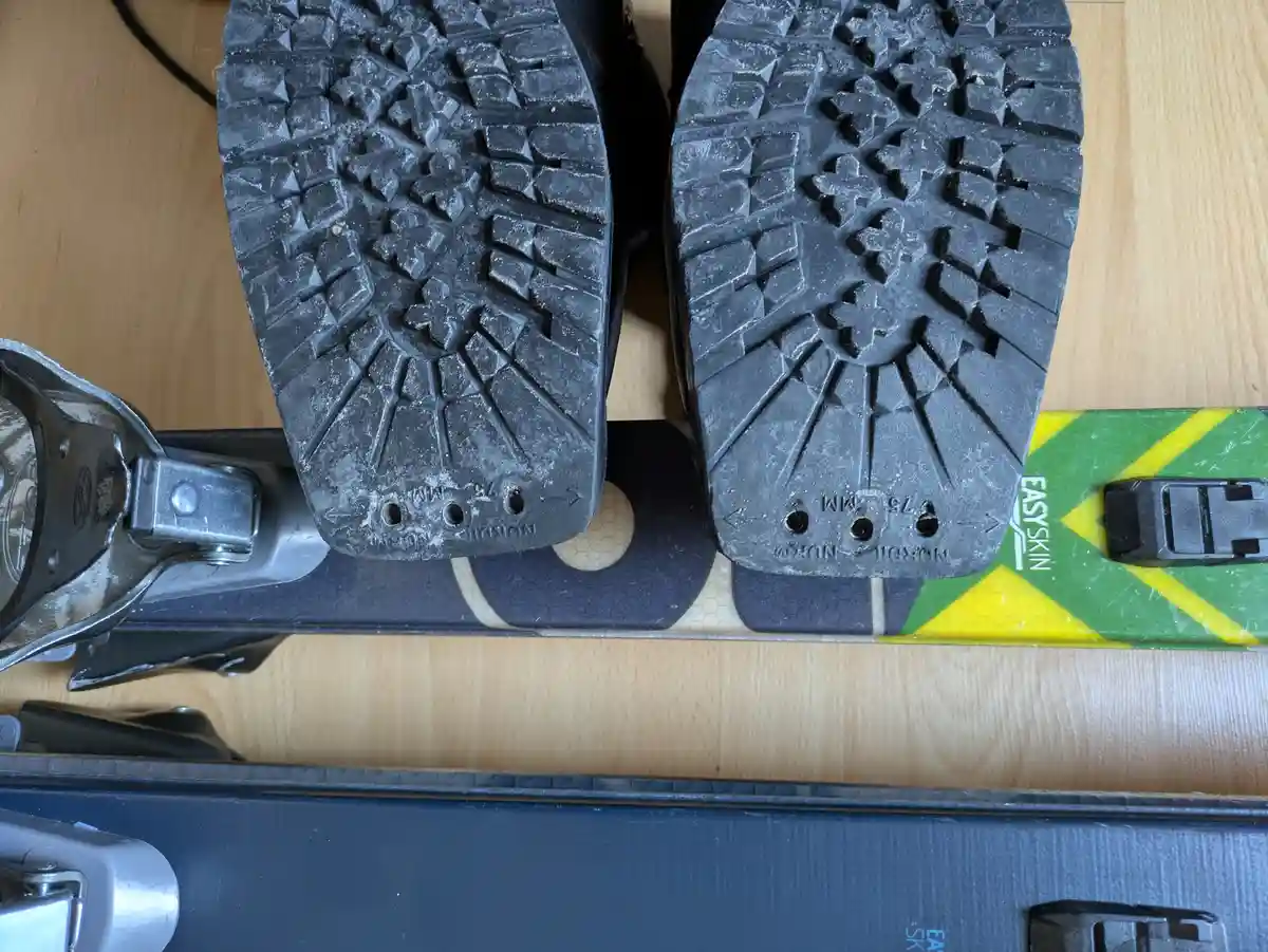 A close-up shot shows the soles of two black ski boots resting on a pair of skis, with the text 'NORDIC NORM 75 MM' visible…