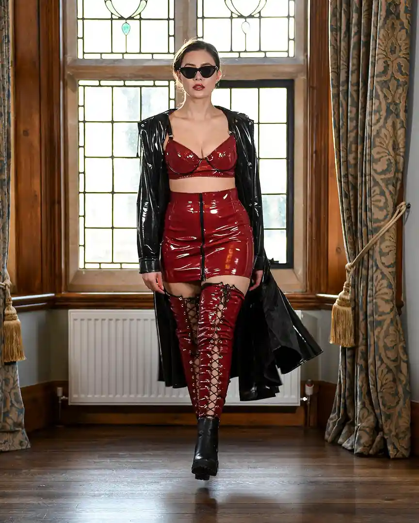 A woman walks confidently in a room with stained glass windows and patterned curtains, wearing a shiny red latex outfit…