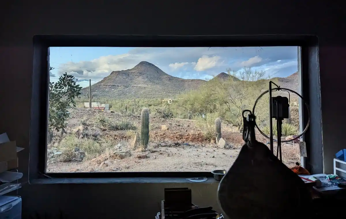 A photo taken from inside a dimly lit room looking out a window at a desert landscape with mountains, cacti, and sparse…