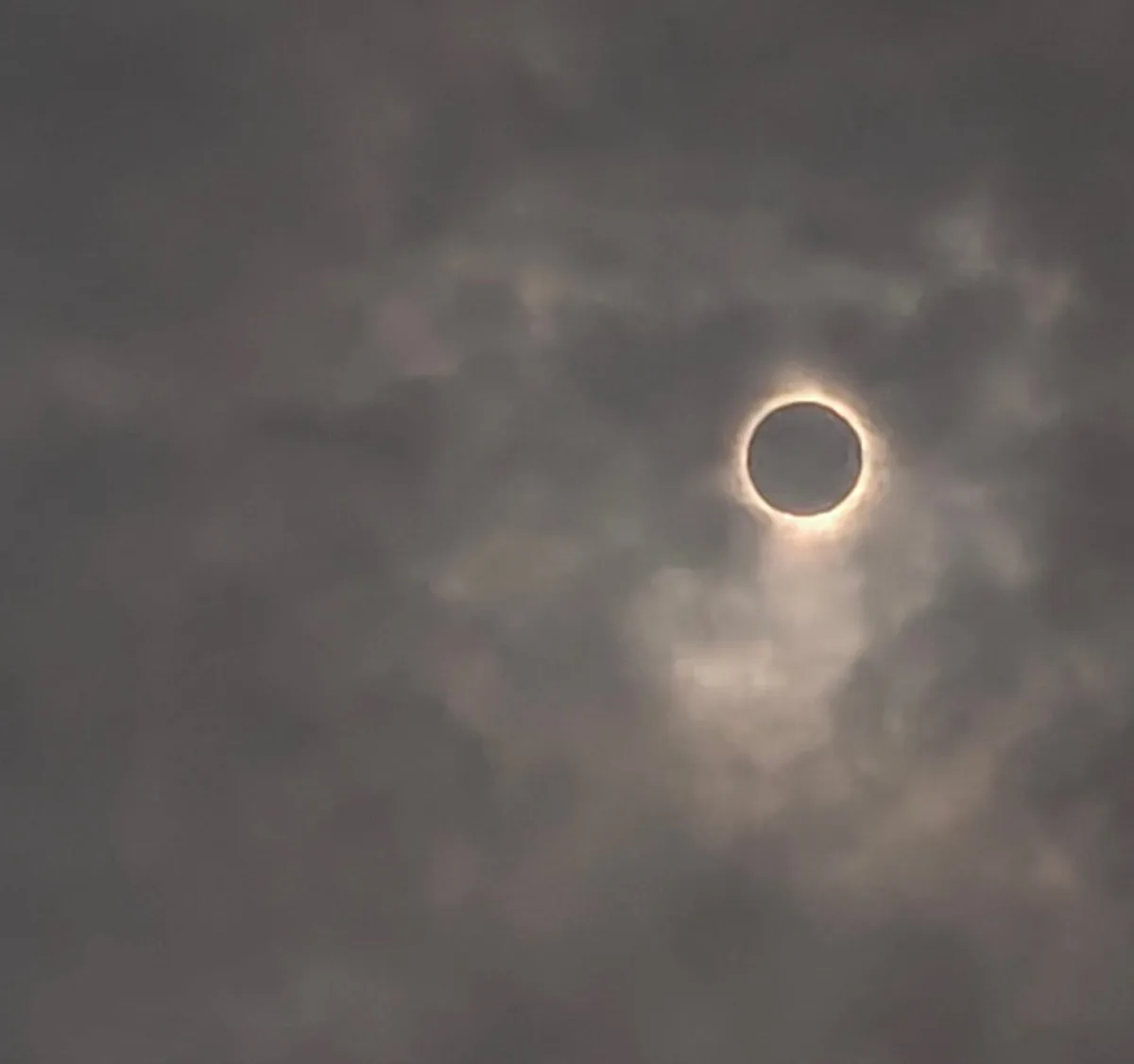 A solar eclipse is visible through a cloudy sky, showing the sun as a dark circle with a glowing ring around it, resembling…