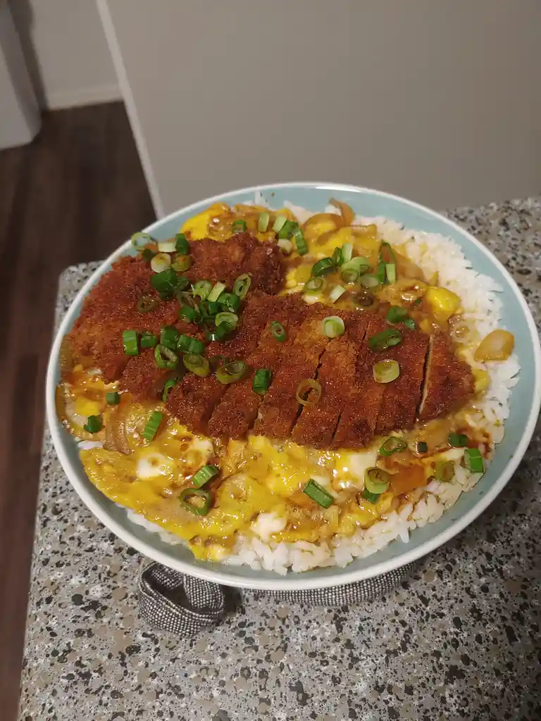 a close-up photo shows a light blue bowl filled with white rice, topped with a sliced breaded pork cutlet, a layer of yellow…