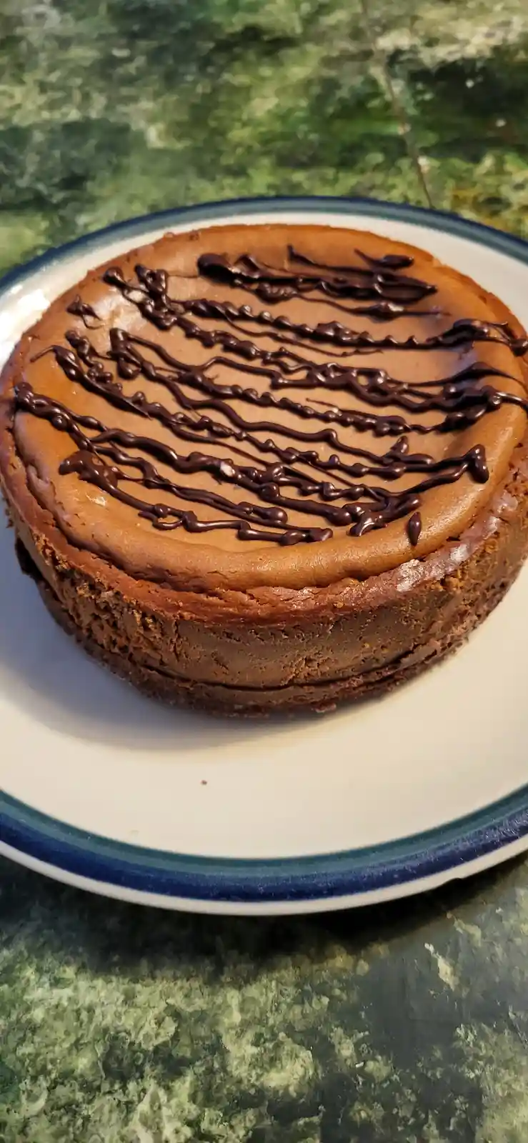 A close-up photo of a round chocolate cheesecake on a white plate with a blue rim, resting on a green marbled countertop…