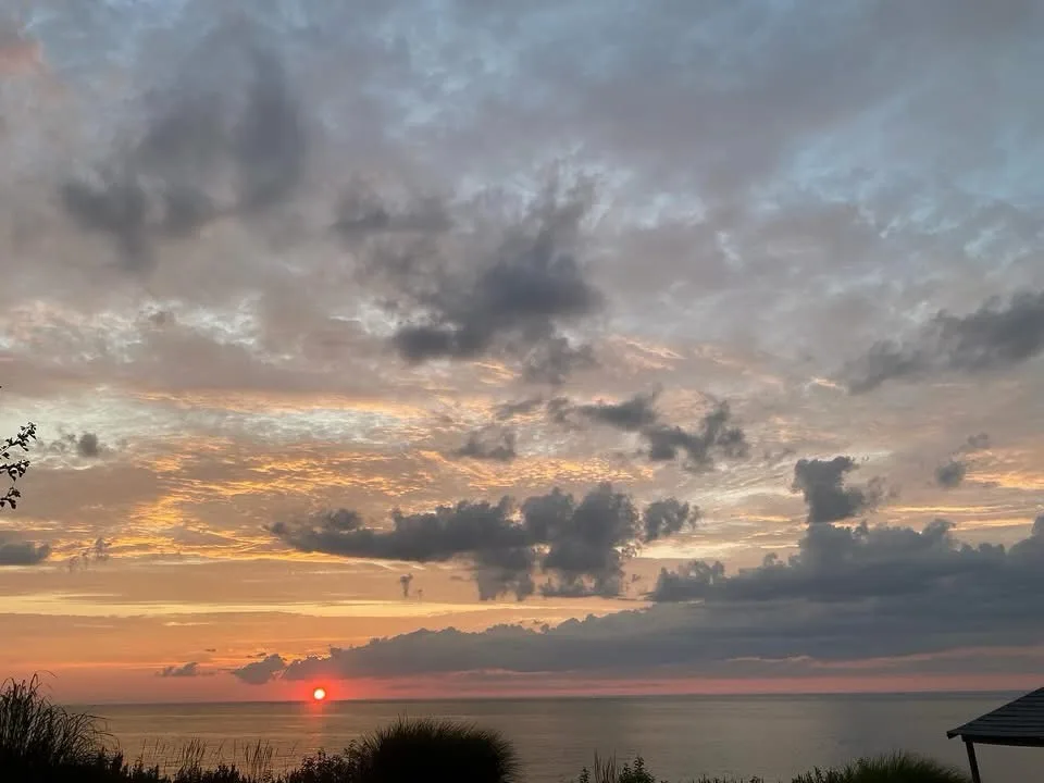 A serene sunset over the ocean with scattered clouds, the sun low on the horizon, and silhouettes of foliage and a roof…
