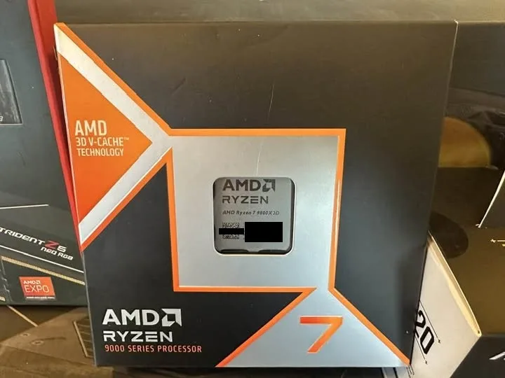 A close-up photo of an AMD Ryzen 7 9800X3D processor box, featuring the AMD logo, '3D V-Cache Technology' text, and a window…