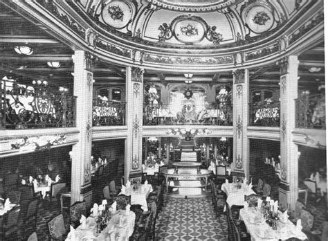 A black-and-white photograph shows the opulent, multi-level dining room of a luxury ship, featuring ornate ceilings…
