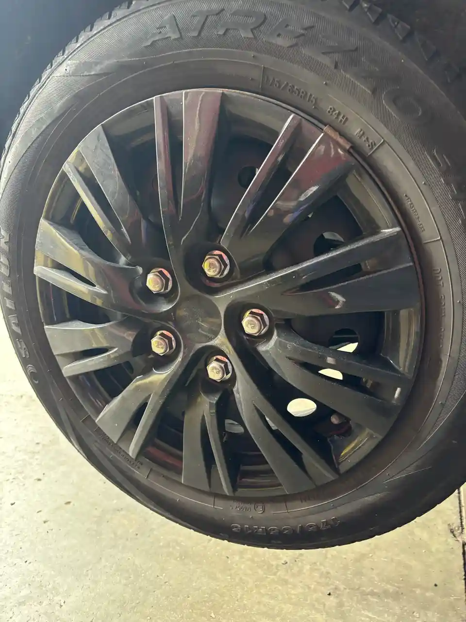 A close-up photo of a shiny, multi-spoke black alloy wheel with a black tire, mounted on a concrete surface. The tire has…