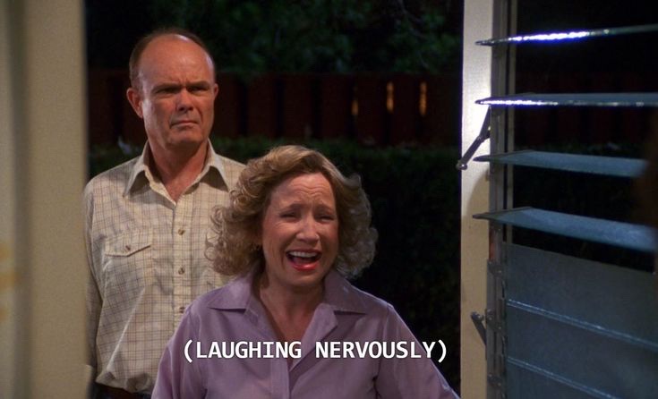 A woman with curly blonde hair in a purple shirt laughs hysterically with her mouth wide open, while a man in a plaid shirt…