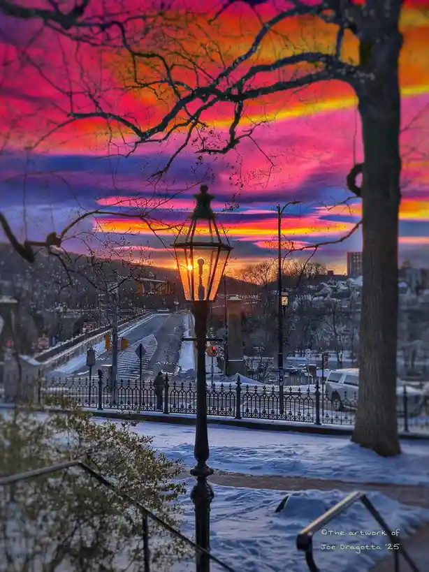 A vibrant winter sunset photo shows a snow-covered street with a glowing ornate streetlamp in the foreground, bare tree…