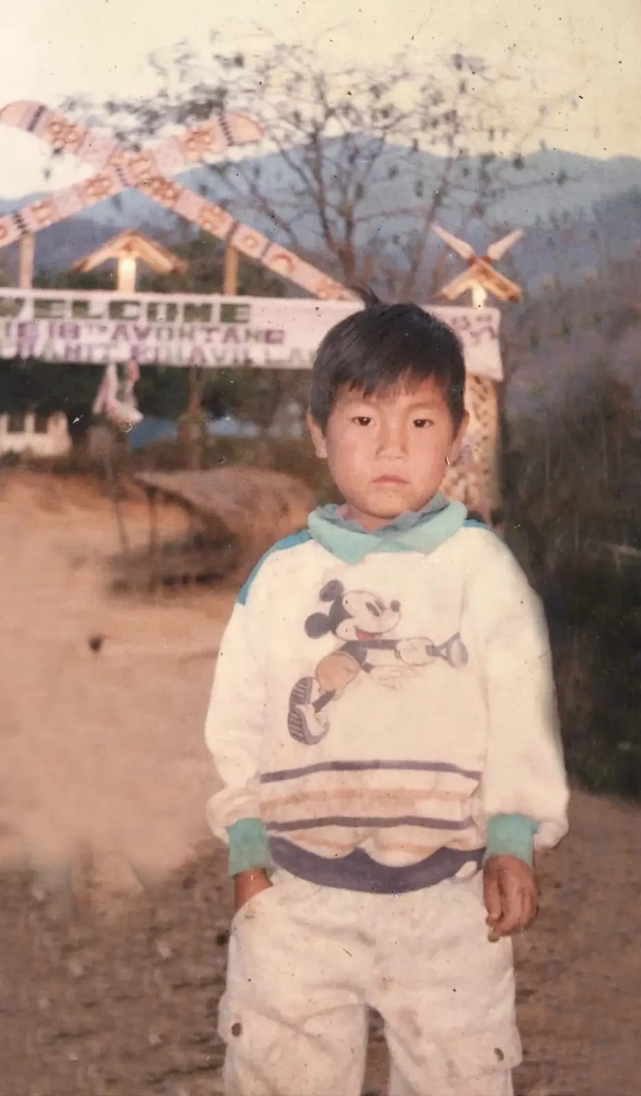 A vintage photo shows a young boy in a Mickey Mouse sweatshirt standing in front of a weathered welcome sign that reads…