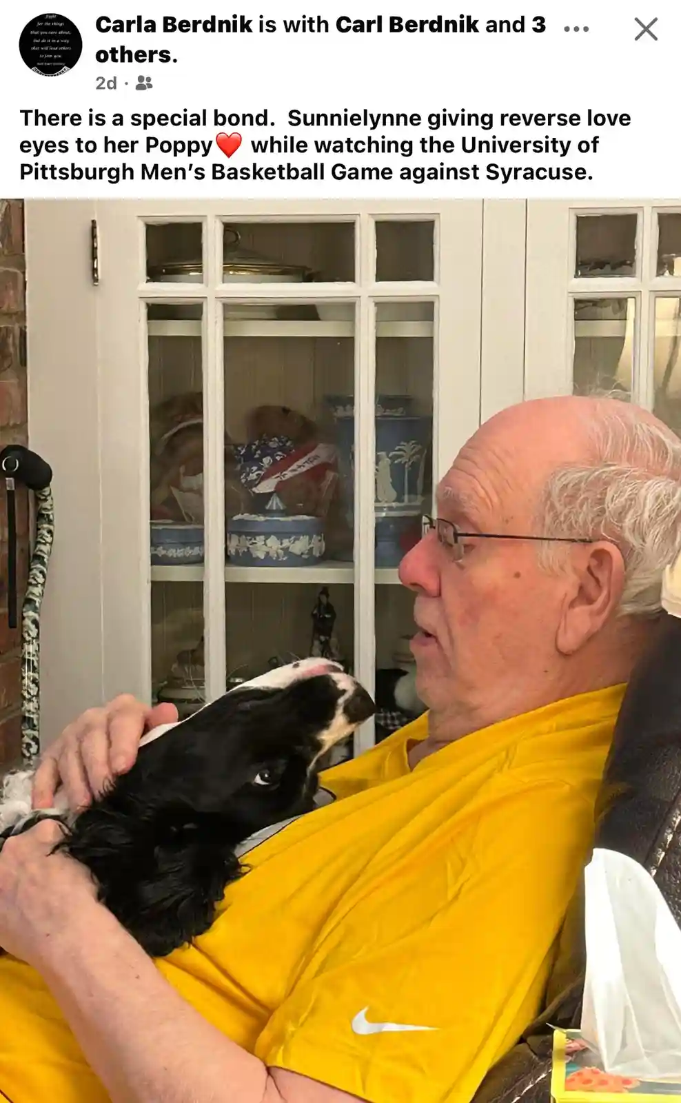 An older man in a yellow shirt sits in a chair, holding a black and white dog on his lap. The dog looks up at him with its…