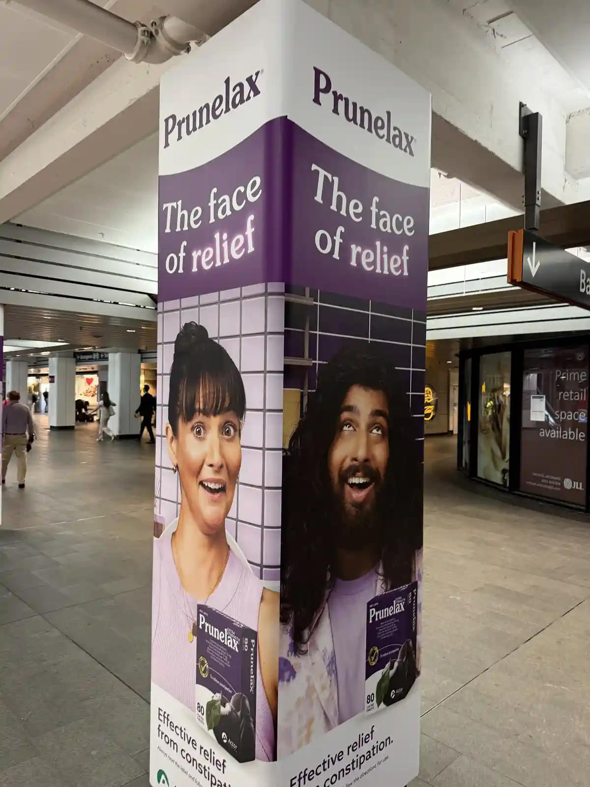 A Prunelax advertisement pillar in a public transit station shows two faces, one of a woman with wide eyes and a surprised…