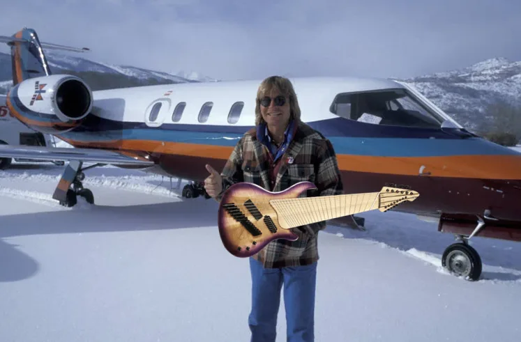 A man in a plaid shirt and sunglasses stands in snow next to a private jet, giving a thumbs-up while holding an unusual…