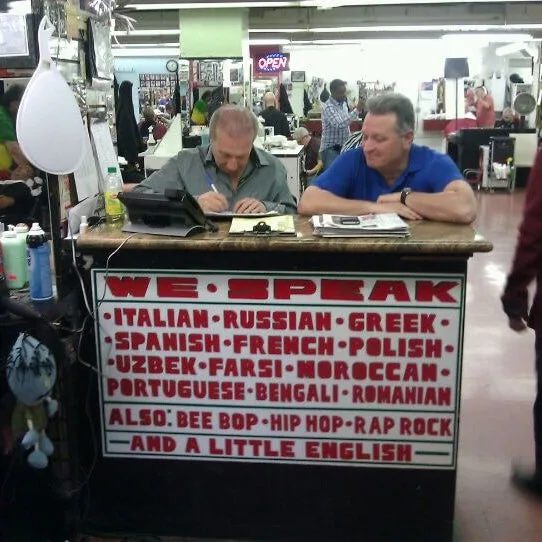 Two men sit at a counter in what appears to be a busy salon or shop, one writing and the other looking on. A large sign on…