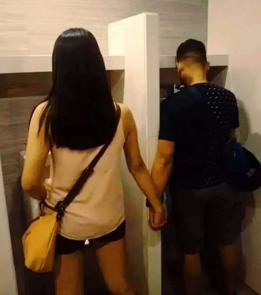 Two people, a woman with long dark hair wearing a beige tank top and carrying a yellow purse, and a man in a dark patterned…