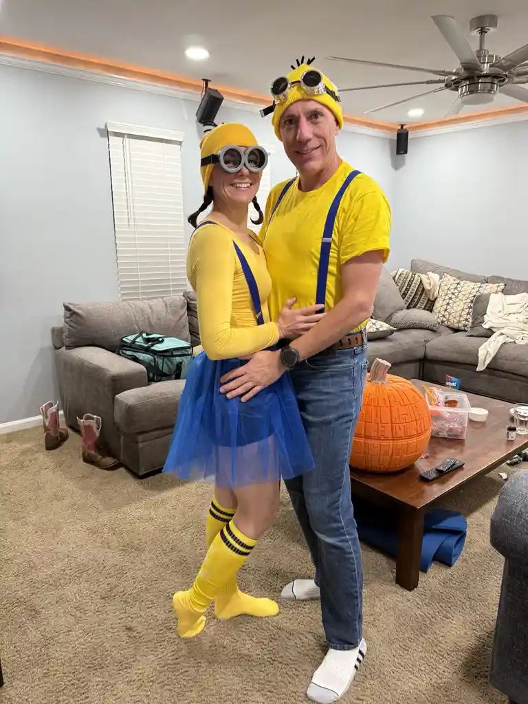 A man and woman pose together in a living room, both dressed as Minions from the Despicable Me franchise. The woman wears a…