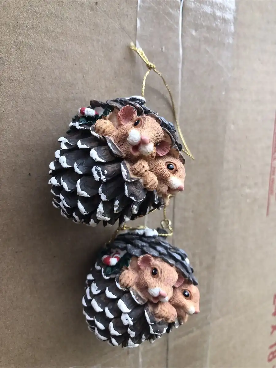 Two identical pinecone-shaped Christmas ornaments hang against a cardboard background, each decorated with miniature brown…