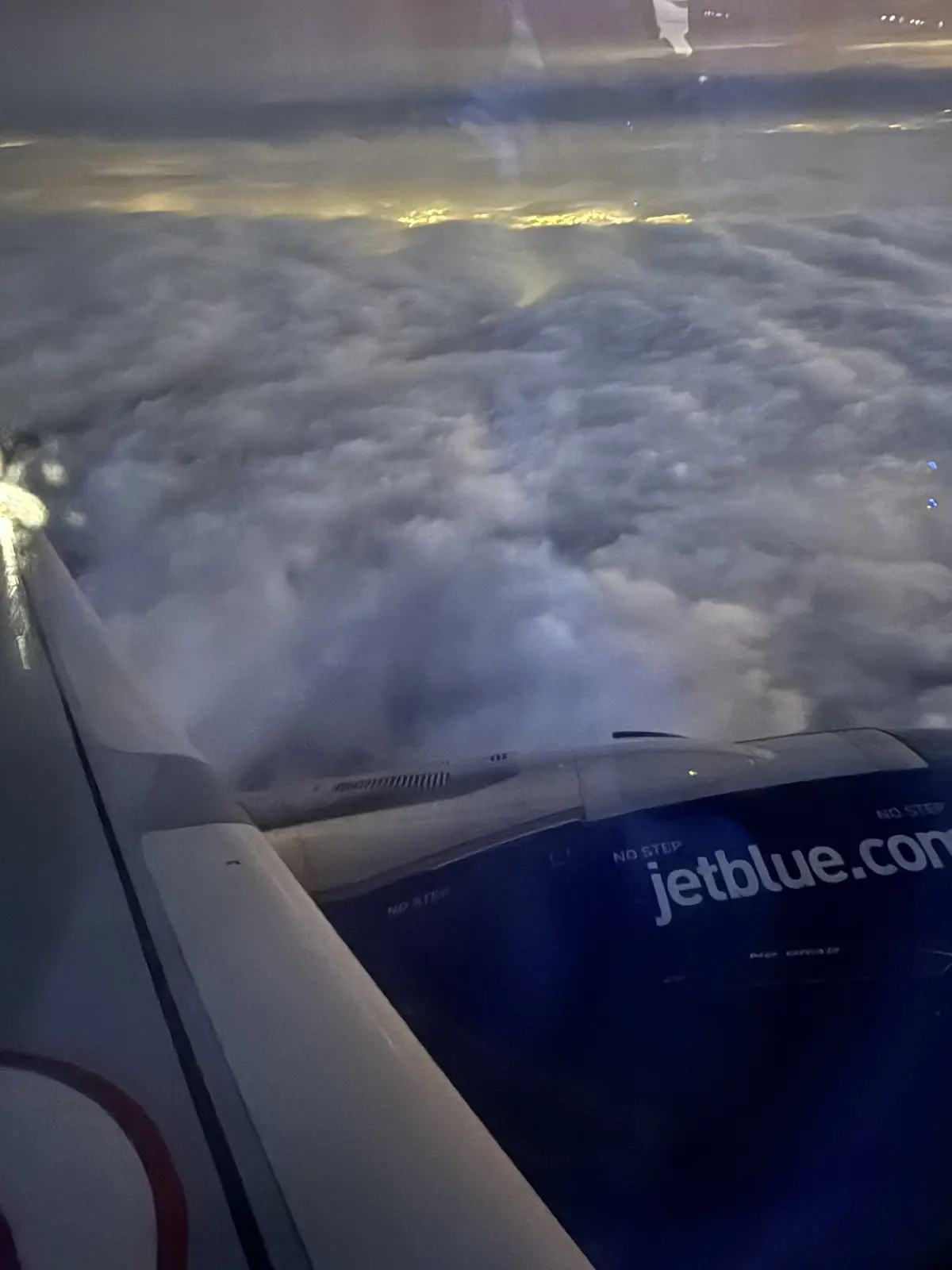 A view from an airplane window showing the wing and engine of a JetBlue aircraft flying above a dense layer of clouds at…