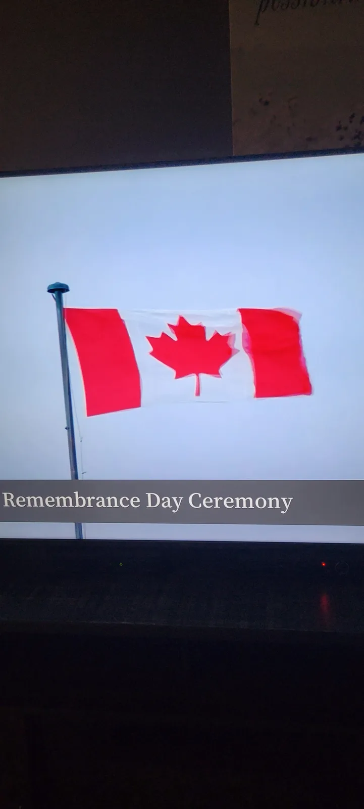 A photograph of a Canadian flag waving on a pole against a pale sky, displayed on a screen. A dark grey banner across the…