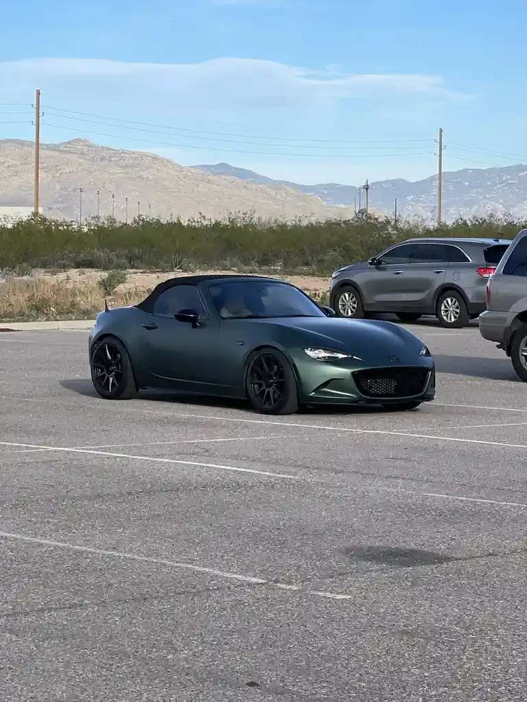 A matte green Mazda MX-5 Miata convertible is parked in an asphalt lot with white lines, surrounded by other cars and desert…