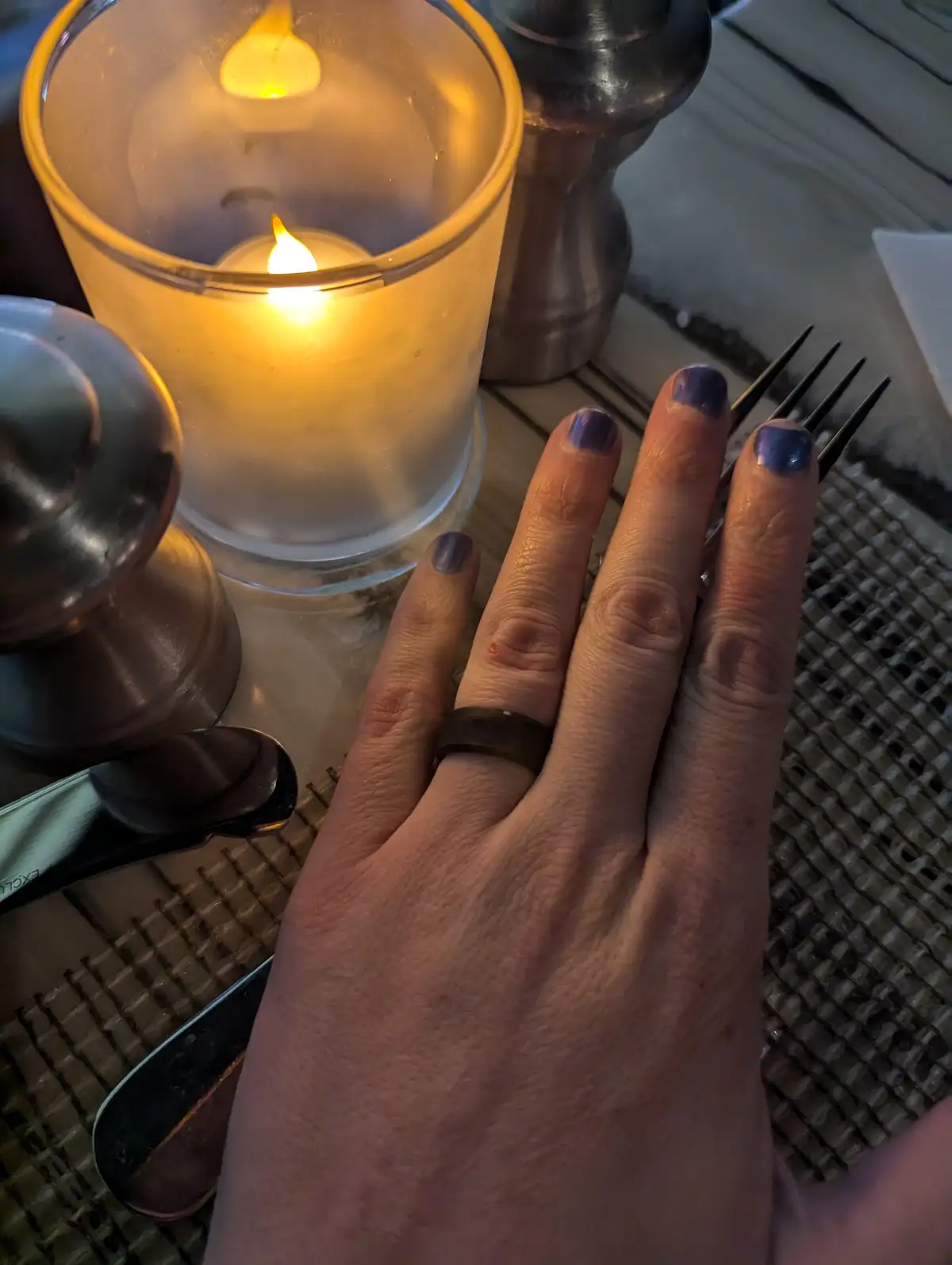 A close-up photo shows a person's hand with purple nail polish resting on a woven placemat next to a lit candle in a glass…