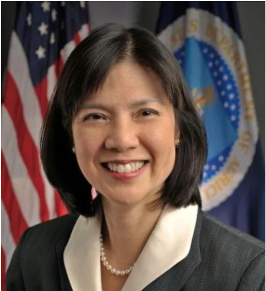 a smiling woman in a dark suit with a white collar and pearl necklace stands in front of an american flag and a blue flag…