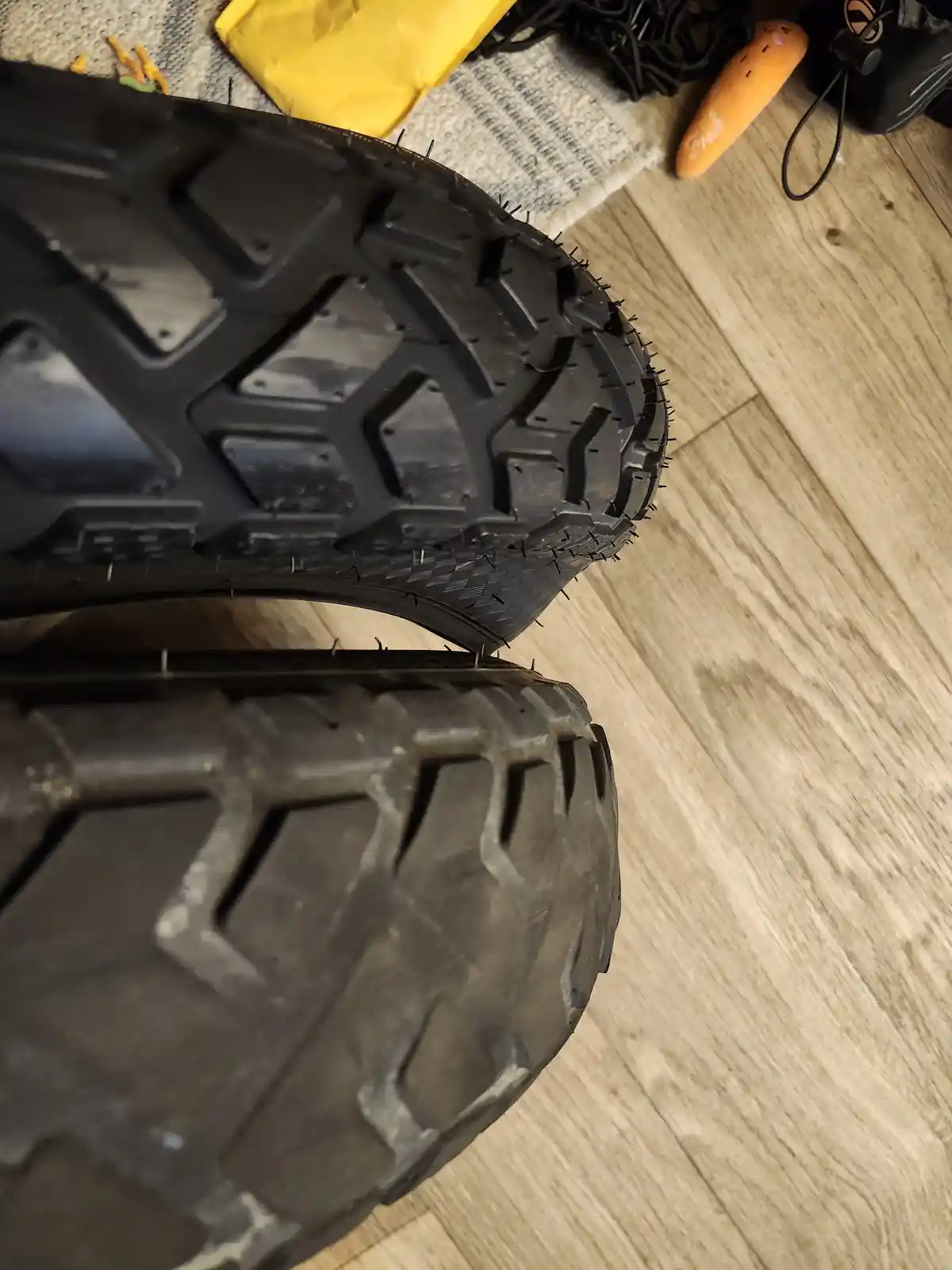 A close-up photo shows two rugged, black off-road tires resting on a light wood floor, with numerous small metal nails…