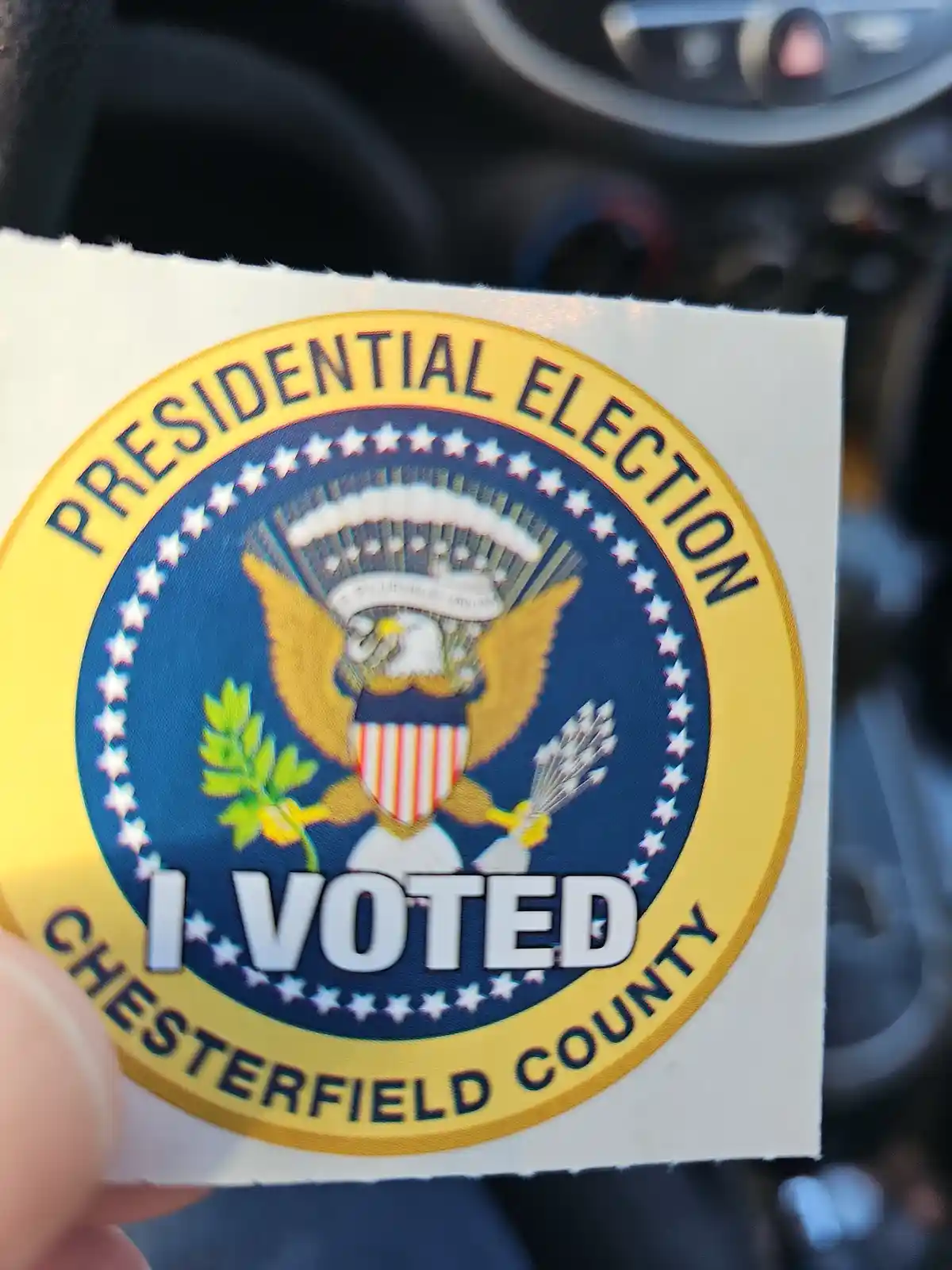 A close-up photo shows a hand holding a circular 'I VOTED' sticker. The sticker features the official seal of the United…