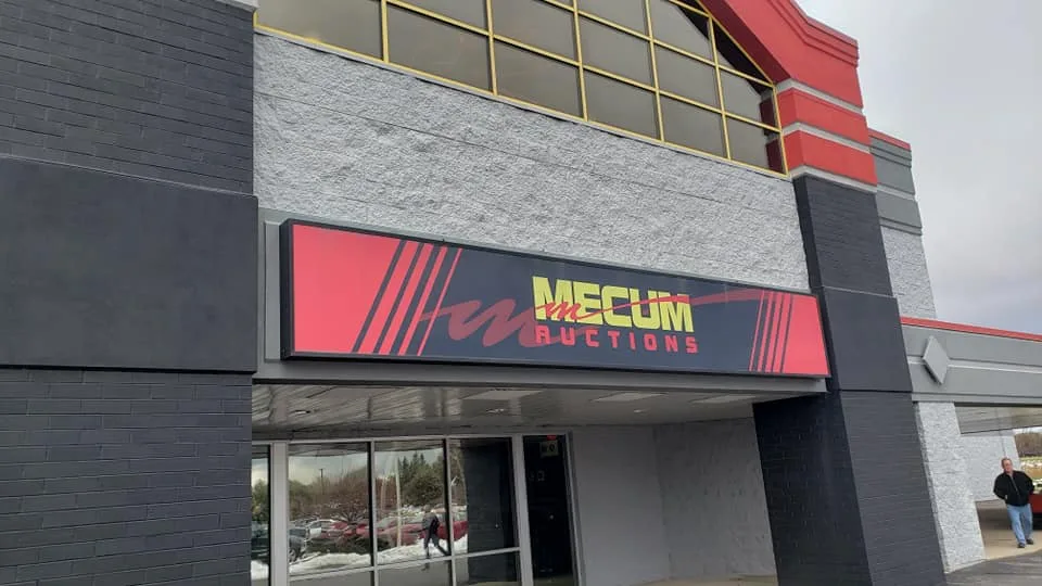 An exterior shot of a modern building with a large sign that reads 'MECUM AUCTIONS' in bold yellow and red lettering against…
