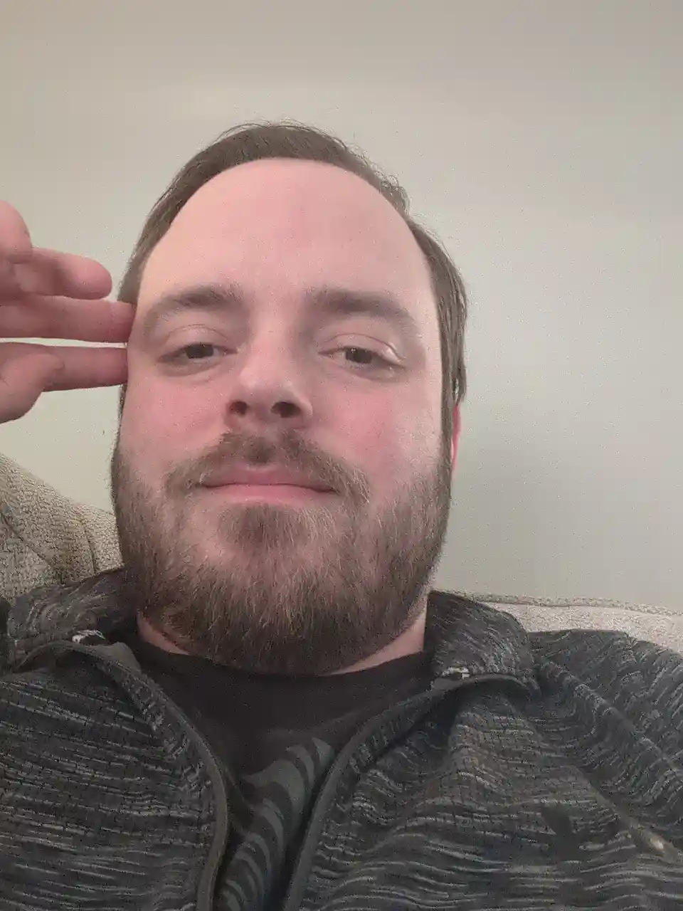 A man with a beard and short hair sits on a couch, looking directly at the camera with a slight smirk while holding his hand…
