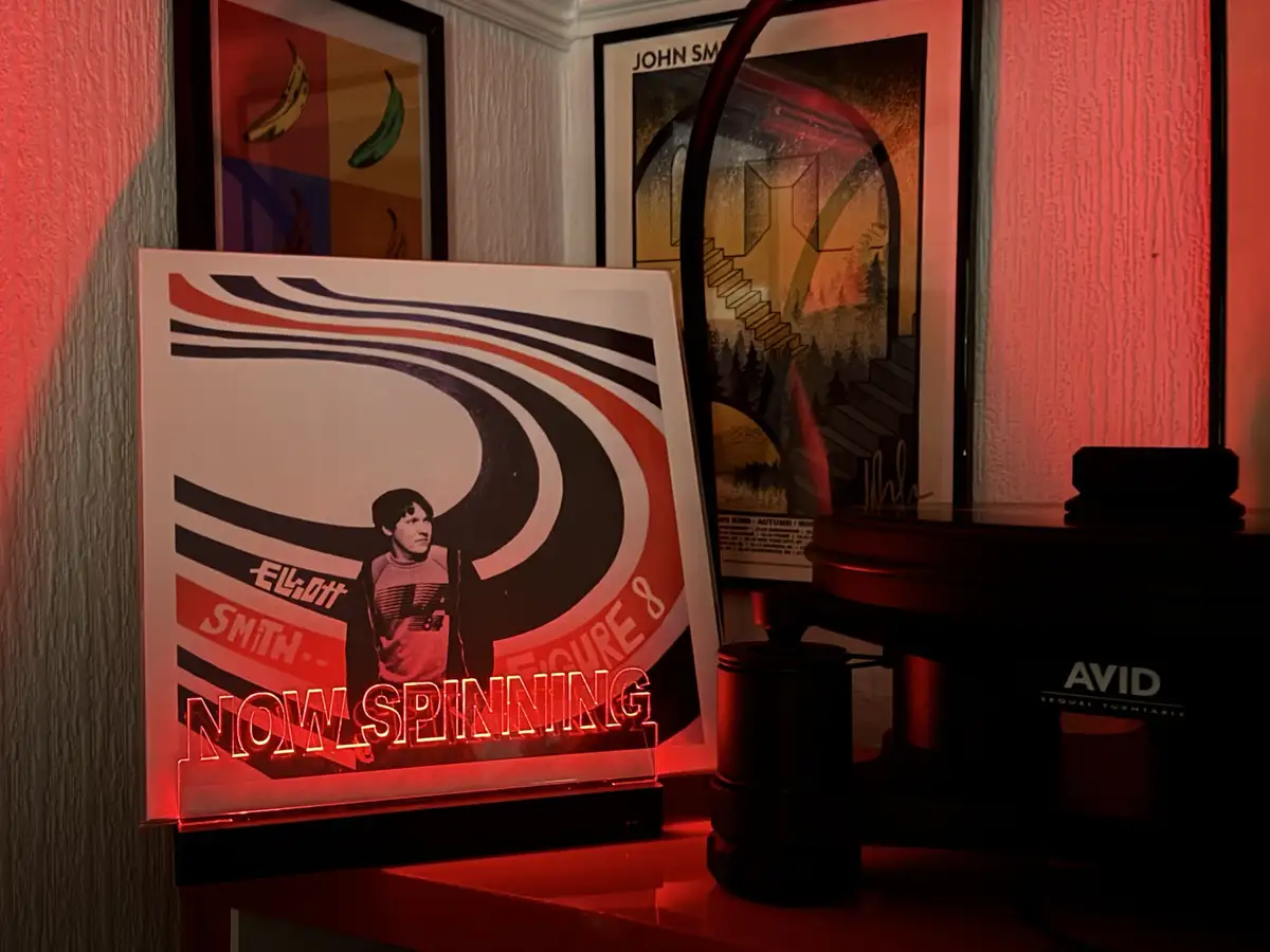 A red-lit neon sign reading 'NOW SPINNING' sits in front of a framed poster of Elliott Smith's album 'Figure 8', placed…