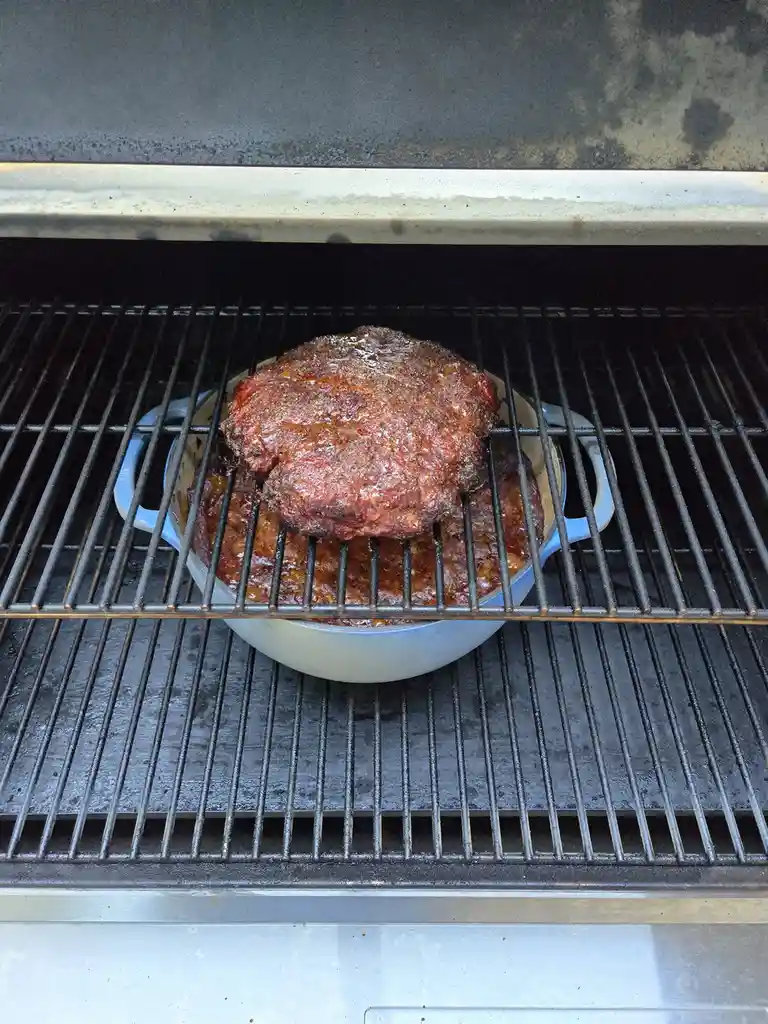A large, glazed meatloaf sits in a light blue Dutch oven on a grill grate inside a barbecue. The meatloaf is browned and…
